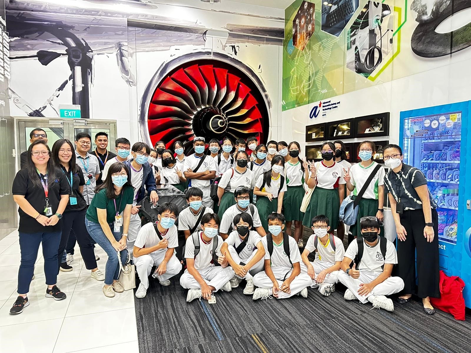 A large group of masked students in white uniforms pose in front of a large red jet engine display indoors.